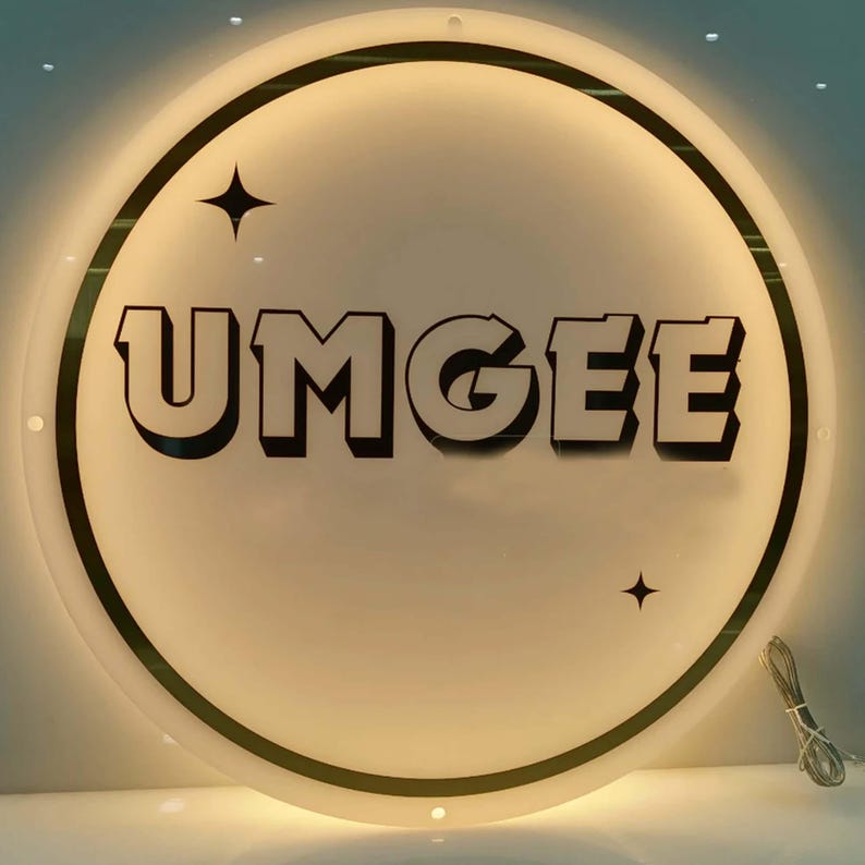 May include: A round, illuminated sign with the word "UMGEE" in bold, black letters. The sign has a white background with a black and gold border. Two black star shapes are on the sign.