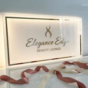 Op de afbeelding: Een wit rechthoekig bord met een gouden rand, met de zilveren woorden "Elegance Edge BEAUTY LOUNGE". Een gouden logo staat boven de tekst. Roze en cr&egrave;mekleurige linten zijn voor het bord geplaatst.