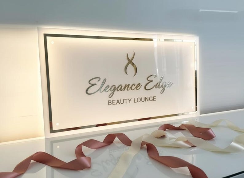 Op de afbeelding: Een wit rechthoekig bord met een gouden rand, met de zilveren woorden "Elegance Edge BEAUTY LOUNGE". Een gouden logo staat boven de tekst. Roze en cr&egrave;mekleurige linten zijn voor het bord geplaatst.
