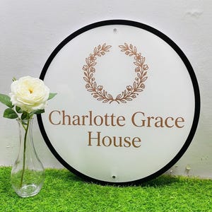 May include: A circular white sign with a thin black border featuring a copper-colored floral wreath graphic above the text Charlotte Grace House in a serif font. The sign has two small mounting holes at the top and bottom center positions.