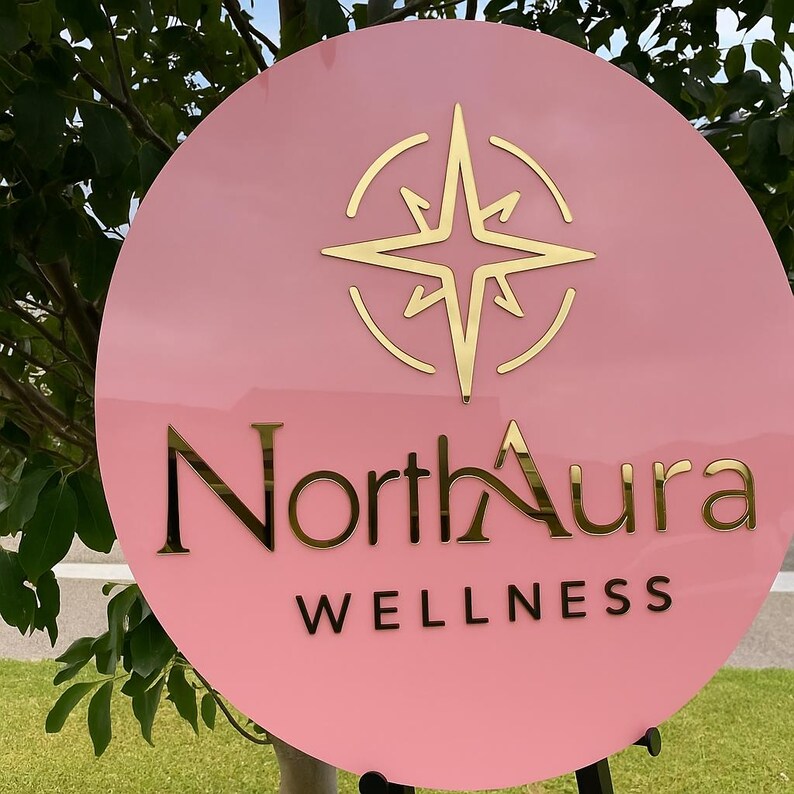 May include: A round, pink sign with gold lettering and a gold compass rose design. The sign reads "North Aura WELLNESS". The sign is displayed outdoors, with green grass and foliage in the background.