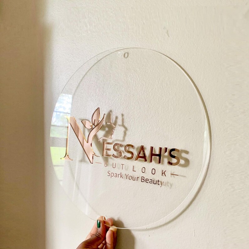 以下が含まれることがあります： NESSAH'S OUTLOOKおよびSpark Your Beautyと書かれたローズゴールドの文字が特徴の円形透明アクリル看板。デザインには、葉のモチーフと横顔のシルエットがあしらわれたスタイリッシュな文字Nが含まれており、上部には小さな穴が開いています。