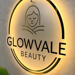 Op de afbeelding: Verlicht cirkelvormig bord voor Glowvale Beauty. Het bord heeft een zwarte omtrek en de woorden "GLOWVALE BEAUTY" in een vetgedrukte, schreefloze letter. Boven de tekst staat een minimalistische lijntekening van een gezicht. Het bord is verlicht met een warme, uitnodigende gloed.