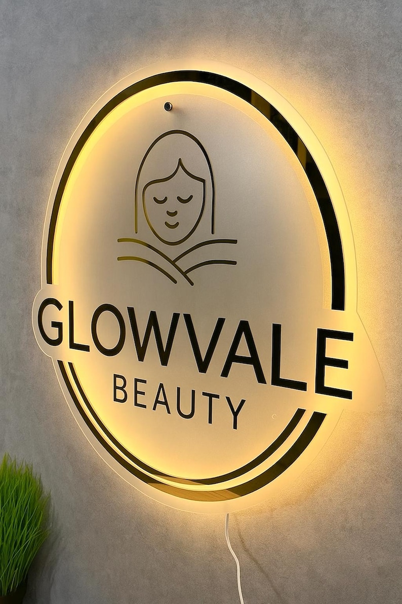 Op de afbeelding: Verlicht cirkelvormig bord voor Glowvale Beauty. Het bord heeft een zwarte omtrek en de woorden "GLOWVALE BEAUTY" in een vetgedrukte, schreefloze letter. Boven de tekst staat een minimalistische lijntekening van een gezicht. Het bord is verlicht met een warme, uitnodigende gloed.