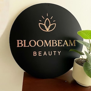 May include: A black circular sign with the words "BLOOMBEAM BEAUTY" in rose gold lettering. Above the text is a rose gold floral design. The sign is mounted on a wall, with a small potted plant visible on the right.