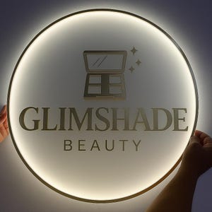 Op de afbeelding: Een ronde, verlichte bewegwijzering met de tekst "GLIMSHADE BEAUTY" in gouden letters. Het bord heeft een gouden oogschaduwpalet-pictogram en steraccenten. Het bord wordt door twee handen vastgehouden.