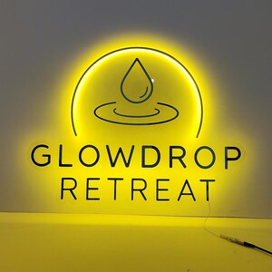 Op de afbeelding: Een heldergeel neonbord met de tekst "GLOWDROP RETREAT" in een schreefloze lettertype. Boven de tekst staat een cirkelvormig ontwerp met een waterdruppel en gebogen lijnen. Het bord licht op tegen een lichtgrijze achtergrond.