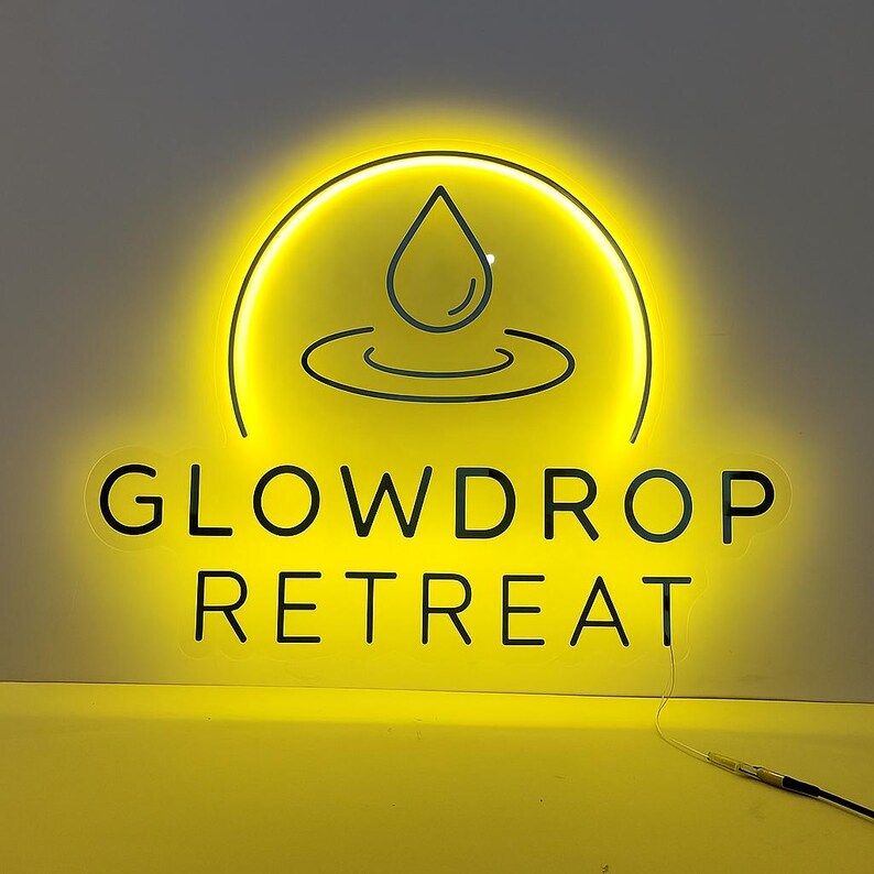 Op de afbeelding: Een heldergeel neonbord met de tekst "GLOWDROP RETREAT" in een schreefloze lettertype. Boven de tekst staat een cirkelvormig ontwerp met een waterdruppel en gebogen lijnen. Het bord licht op tegen een lichtgrijze achtergrond.