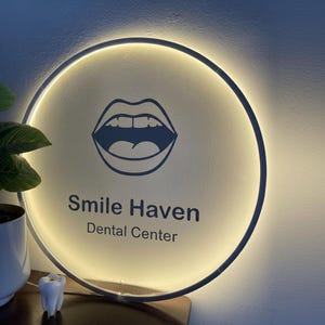 Op de afbeelding: Rond verlicht bord voor een tandartspraktijk. Het bord heeft een donkerblauwe rand en een witte binnenkant met de woorden "Smile Haven Dental Center" in donkerblauw. Een afbeelding van open lippen staat boven de tekst.