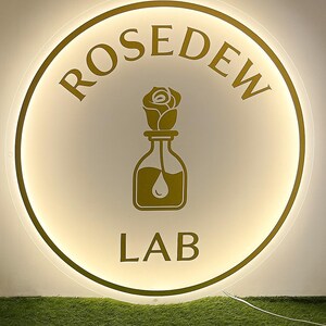 Op de afbeelding: Een rond, verlicht bord met de woorden "ROSED EW LAB" in goud. Het bord heeft een gouden roos en fles afbeelding. Het bord staat tegen een witte achtergrond en is omgeven door een gouden rand.