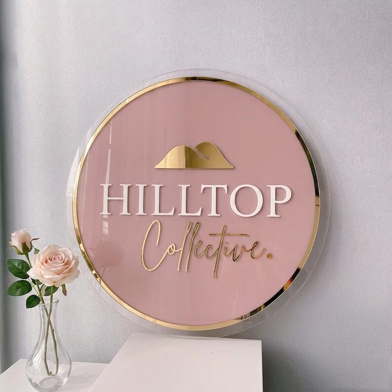 May include: A circular sign with a dusty pink background and a gold metallic border. The sign features a gold mountain icon, the white text HILLTOP, and the gold cursive text Collective. A clear acrylic layer frames the entire circular design.