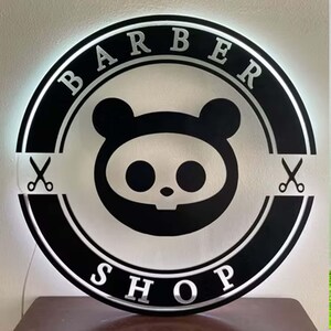 May include: A round, black and white Barber Shop sign with the words "BARBER SHOP" in silver lettering. The sign features a cartoon panda-like skull design in the center and scissors on either side.