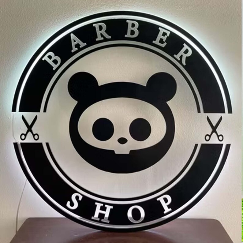 May include: A round, black and white Barber Shop sign with the words "BARBER SHOP" in silver lettering. The sign features a cartoon panda-like skull design in the center and scissors on either side.