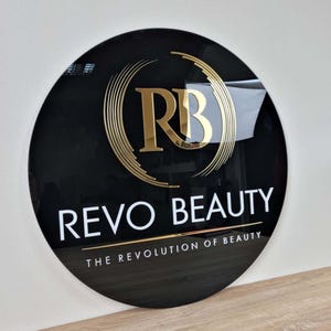 May include: A black circular sign with gold lettering. The sign reads "REVO BEAUTY" and "THE REVOLUTION OF BEAUTY". The logo is a gold "RB" inside a circle of gold lines.
