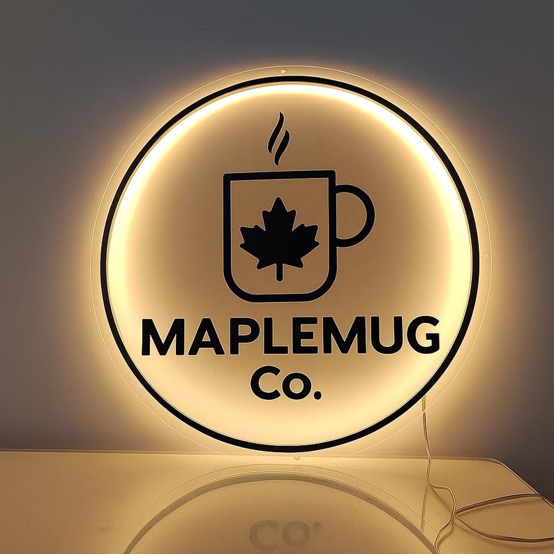 Op de afbeelding: Rond verlicht bord met de tekst "MAPLEMUG Co." in een vet, zwart lettertype. Het bord heeft een zwarte afbeelding van een koffiemok met een esdoornblad erin en stoom die van bovenaf opstijgt. Het bord is omgeven door een gloeiend, warm licht.