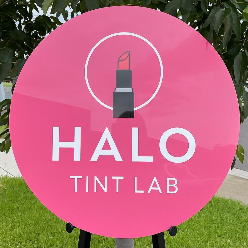 May include: A round, pink sign with the words "HALO TINT LAB" in white. Above the text is a white circle with a lipstick icon. The sign is mounted on a black stand, with green grass and foliage in the background.