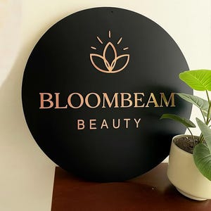 May include: A round black sign with the words "BLOOMBEAM BEAUTY" in rose gold lettering. Above the text is a rose gold floral design. The sign is mounted on a wall, with a small potted plant to the right.