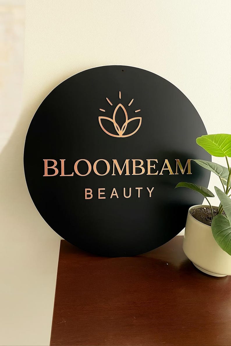 May include: A round black sign with the words "BLOOMBEAM BEAUTY" in rose gold lettering. Above the text is a rose gold floral design. The sign is mounted on a wall, with a small potted plant to the right.