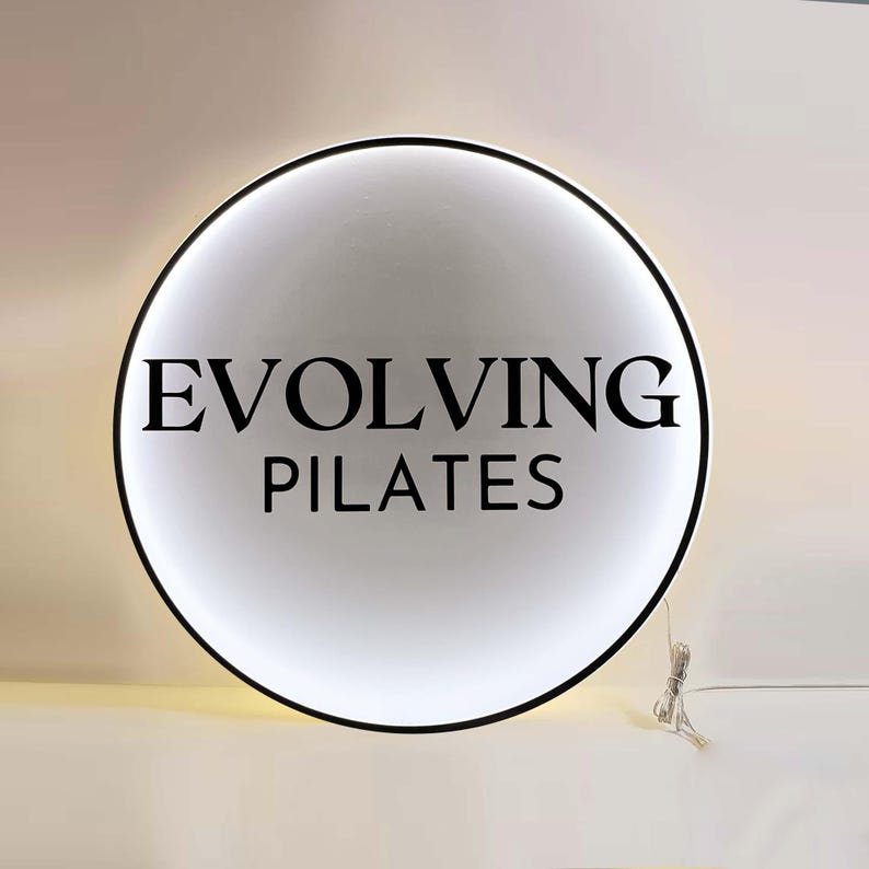 May include: A round, illuminated sign with the words "EVOLVING PILATES" in black, centered on a white background. The sign has a black border and a power cord extending from the bottom.