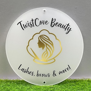 May include: A round, white sign with the text "Twist Cove Beauty" in black script. The sign features a gold illustration of a woman's profile within a gold cloud-like shape. The text "Lashes, brows & more!" is also included.