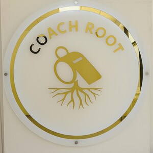 May include: A round sign with the words "COACH ROOT" in gold lettering. The sign features a gold whistle graphic above a gold root design, all set against a white background with a gold border.