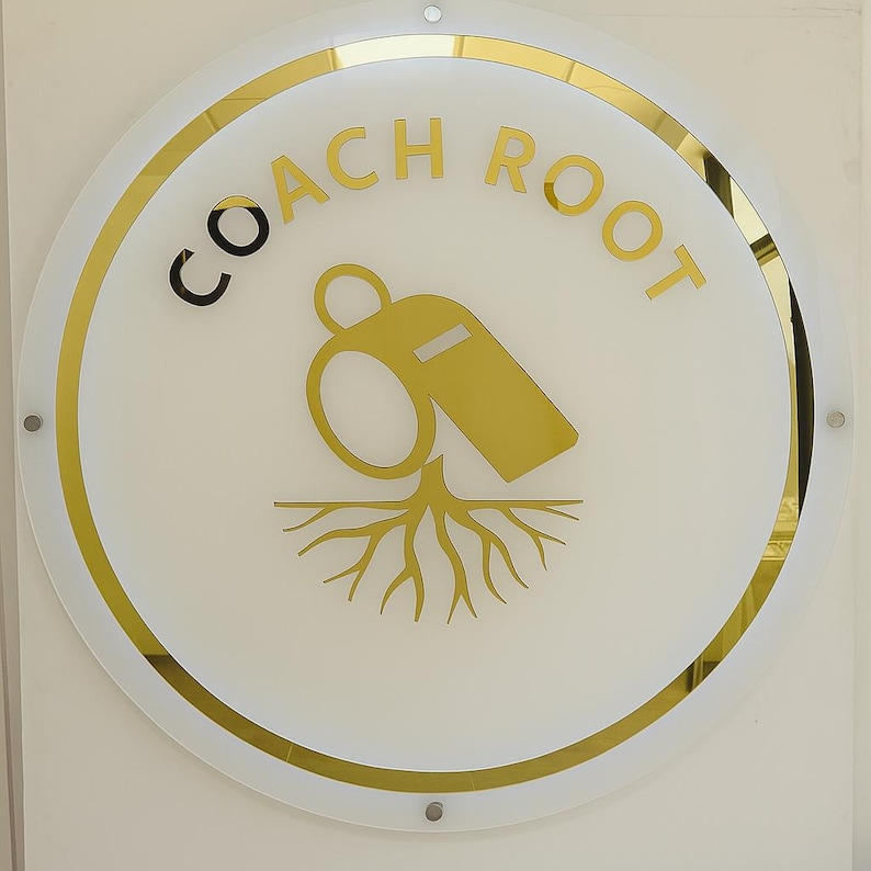 May include: A round sign with the words "COACH ROOT" in gold lettering. The sign features a gold whistle graphic above a gold root design, all set against a white background with a gold border.