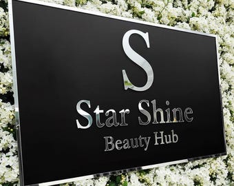 Può includere: Un cartello rettangolare nero con bordo e scritte a specchio argentate. Il cartello recita "Star Shine Beauty Hub" in un carattere a specchio. Lo sfondo è un muro di fiori bianchi.