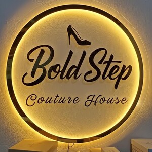 Op de afbeelding: Een rond verlicht bord met de tekst "Bold Step Couture House" in zwart script. Een zwarte afbeelding van een high-heelschoen staat boven de tekst. Het bord heeft een gouden rand en een warme, uitnodigende gloed.