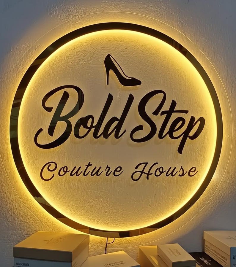 Op de afbeelding: Een rond verlicht bord met de tekst "Bold Step Couture House" in zwart script. Een zwarte afbeelding van een high-heelschoen staat boven de tekst. Het bord heeft een gouden rand en een warme, uitnodigende gloed.
