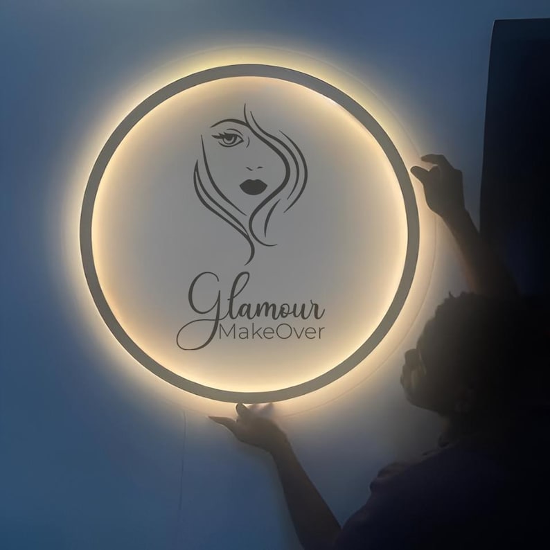 May include: A circular wall sign featuring a backlit white surface with a black line art illustration of a face and the text Glamour MakeOver. The sign emits a soft warm glow from behind its circular frame, creating a halo effect.