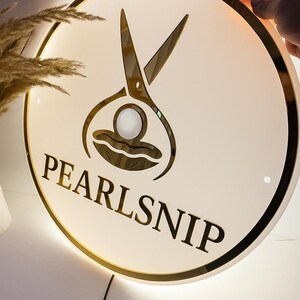 Op de afbeelding: Ronde, verlichte bewegwijzering met een gouden rand en de tekst "PEARLSNIP". De bewegwijzering heeft een gouden schaargrafiek boven de tekst. De bewegwijzering is wit met een gouden rand en een zwart netsnoer.