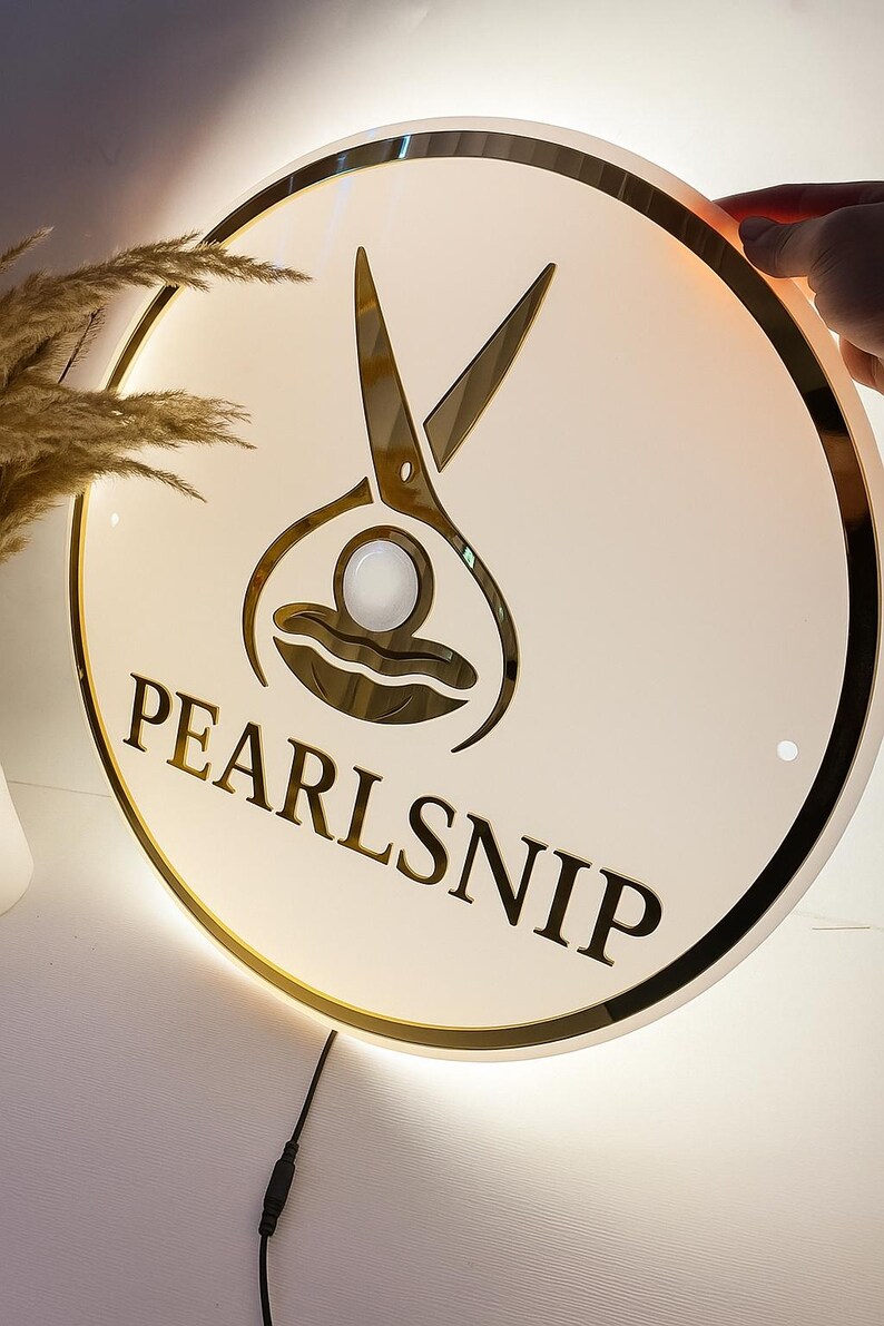 Op de afbeelding: Ronde, verlichte bewegwijzering met een gouden rand en de tekst "PEARLSNIP". De bewegwijzering heeft een gouden schaargrafiek boven de tekst. De bewegwijzering is wit met een gouden rand en een zwart netsnoer.