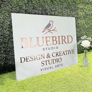 May include: A white rectangular sign featuring a blue bird icon perched on a branch above the rose gold text BLUEBIRD STUDIO DESIGN & CREATIVE STUDIO VISUAL ARTS. A clear glass vase with a single white rose stands next to the sign.