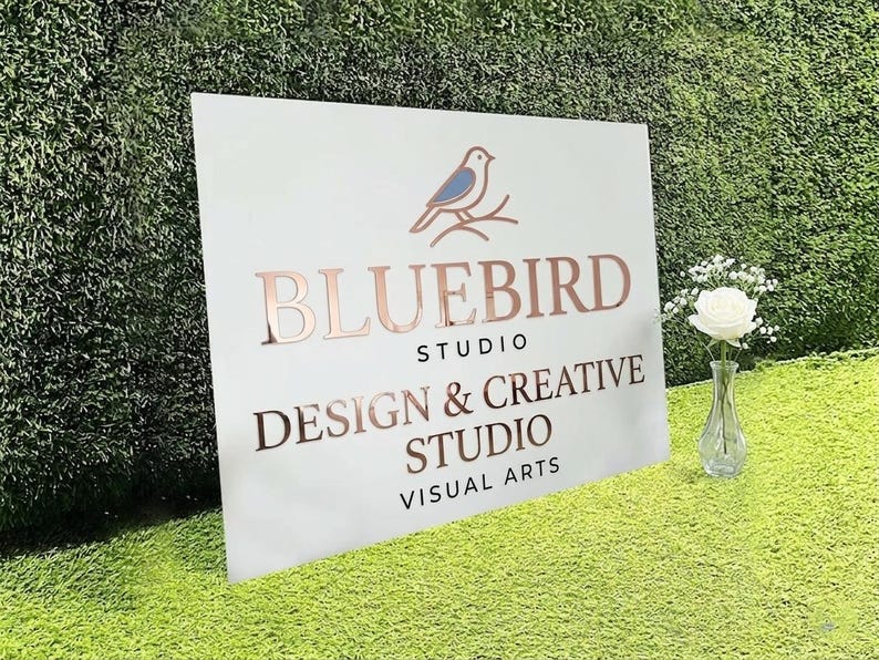 May include: A white rectangular sign featuring a blue bird icon perched on a branch above the rose gold text BLUEBIRD STUDIO DESIGN & CREATIVE STUDIO VISUAL ARTS. A clear glass vase with a single white rose stands next to the sign.