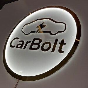 May include: A round, illuminated sign with a gold-colored trim. The sign features a car silhouette with a lightning bolt and the text "CarBolt" in a bold font. The background is white with a bright, glowing effect.