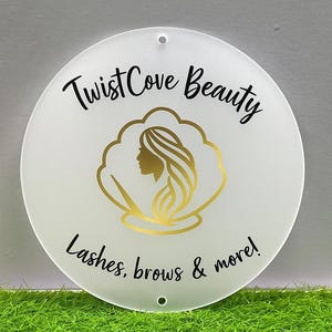 May include: A round, white sign with the text "Twist Cove Beauty" in black script. The sign features a gold illustration of a woman's profile within a gold cloud-like shape. The text "Lashes, brows & more!" is also included.