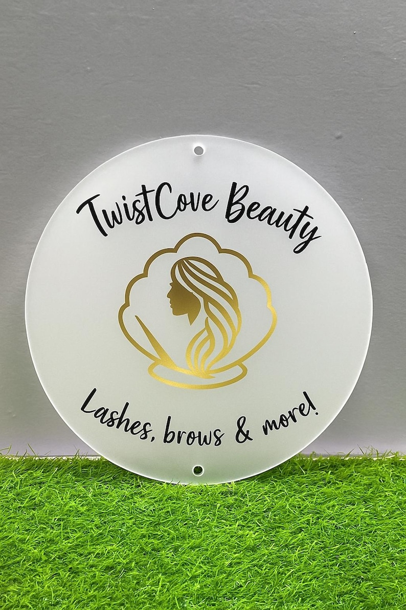 May include: A round, white sign with the text "Twist Cove Beauty" in black script. The sign features a gold illustration of a woman's profile within a gold cloud-like shape. The text "Lashes, brows & more!" is also included.