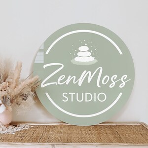 May include: A round, sage green sign with the text "ZenMoss STUDIO" in white script. The sign features a white graphic of stacked stones and is displayed near dried flowers in a vase.