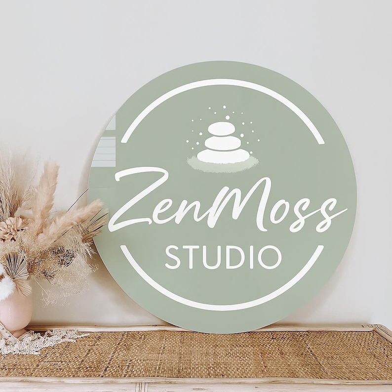 May include: A round, sage green sign with the text "ZenMoss STUDIO" in white script. The sign features a white graphic of stacked stones and is displayed near dried flowers in a vase.