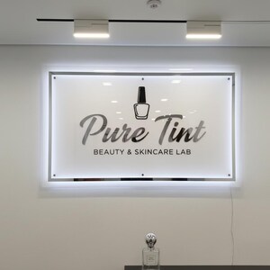 Op de afbeelding: Een rechthoekig verlicht bord met de tekst "Pure Tint BEAUTY & SKINCARE LAB" en een nagellakfles-icoon. Een parfumfles staat eronder op een donkere ondergrond.