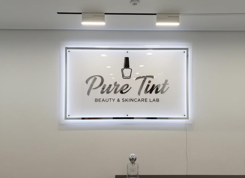 Op de afbeelding: Een rechthoekig verlicht bord met de tekst "Pure Tint BEAUTY & SKINCARE LAB" en een nagellakfles-icoon. Een parfumfles staat eronder op een donkere ondergrond.