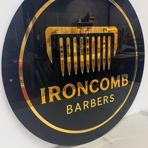 May include: A round black sign with gold trim and lettering. The sign features a gold comb graphic and the words "IRONCOMB BARBERS" in gold. The sign is mounted on a white wall.