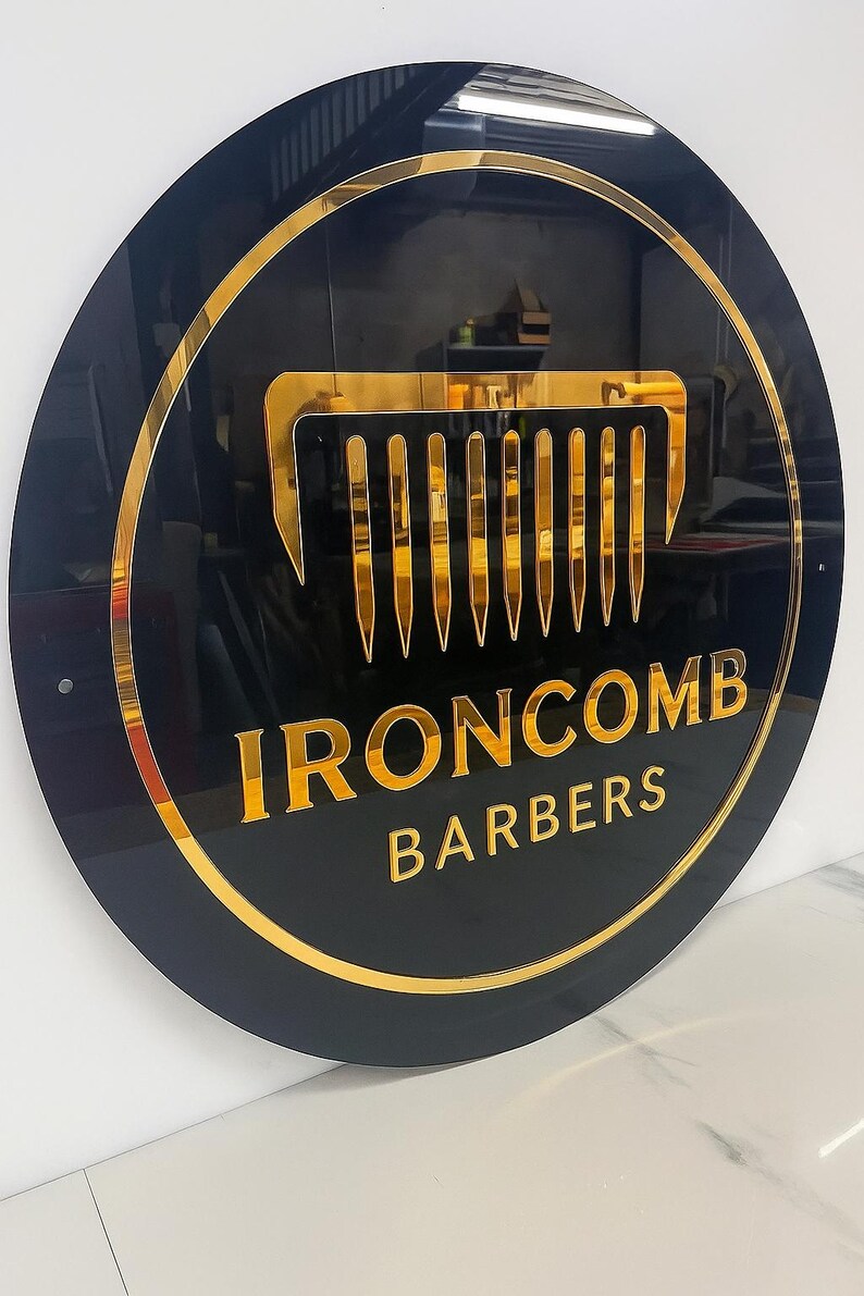 May include: A round black sign with gold trim and lettering. The sign features a gold comb graphic and the words "IRONCOMB BARBERS" in gold. The sign is mounted on a white wall.