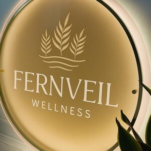 Op de afbeelding: Een ronde, verlichte bewegwijzering met de tekst "FERNVEIL WELLNESS" in het wit. Het bord heeft een goudkleurig midden met een bladerontwerp en een witte rand. Het is waarschijnlijk voor een wellnesscentrum.