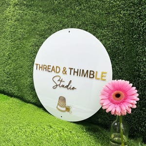 May include: A circular white sign featuring the gold text THREAD & THIMBLE STUDIO, with a gold thimble icon below the lettering. The sign has two small holes at the top and bottom and is positioned next to a pink gerbera daisy in a glass vase.