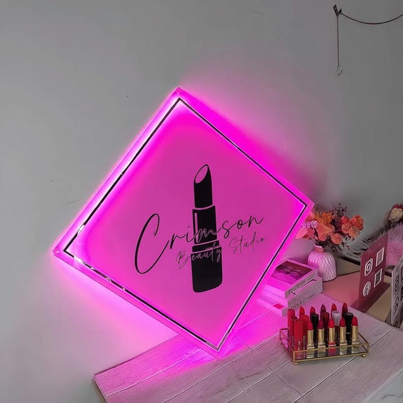 Op de afbeelding: Een roze, verlicht bord met de tekst "Crimson Beauty Studio" en een lippenstiftafbeelding. Het ruitvormige bord heeft een zilveren rand. Een display met lippenstiften staat eronder.