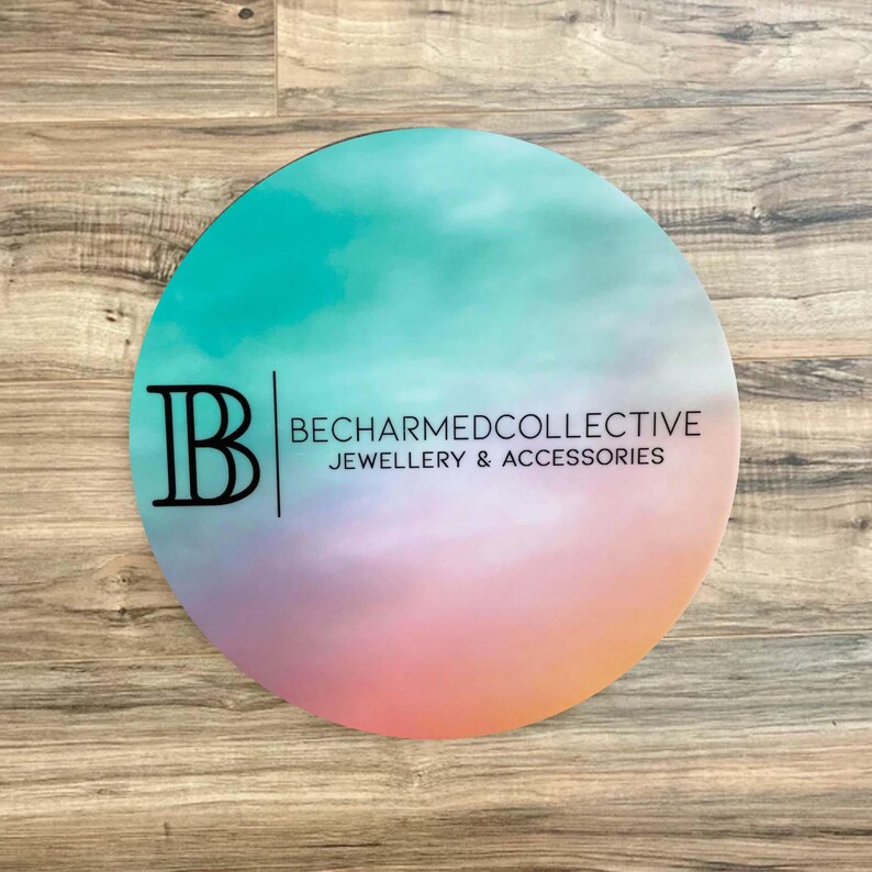 May include: A round, colorful sign with a gradient of teal, pink, and orange. The sign has a black letter "B" and the text "BECHARMEDCOLLECTIVE JEWELLERY & ACCESSORIES".
