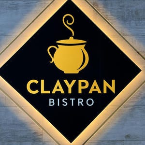 Op de afbeelding: Verlicht bord voor Claypan Bistro. Het bord heeft een zwarte ruitvorm met een gouden omtrek en een gouden afbeelding van een pot met stoom. De woorden "CLAYPAN" en "BISTRO" worden in goud en wit weergegeven.