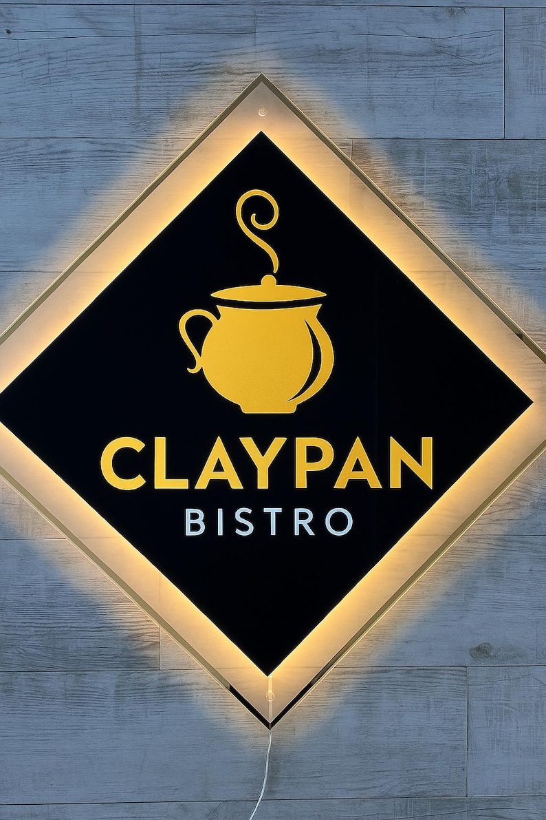 Op de afbeelding: Verlicht bord voor Claypan Bistro. Het bord heeft een zwarte ruitvorm met een gouden omtrek en een gouden afbeelding van een pot met stoom. De woorden "CLAYPAN" en "BISTRO" worden in goud en wit weergegeven.