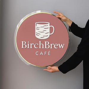 May include: A round sign for BirchBrew Caf&eacute;, featuring a white coffee cup graphic and text on a dusty rose background. The sign has a gold border and is held by hands.